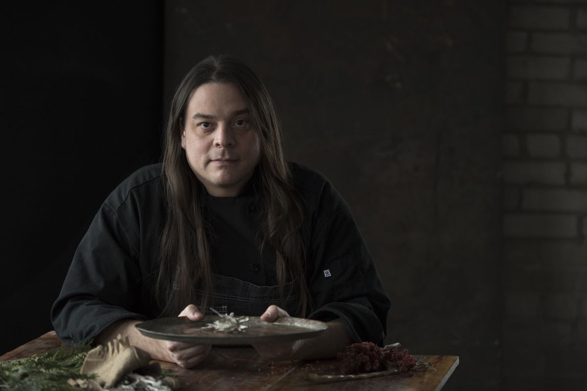 Man with long hair sitting behind table with a dark black background.