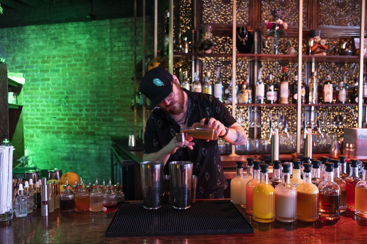 A bartender with glasses and a beard in a black hat and black shirt pouring into a shot glass with shelving in the background and many bottles in front at Midnight Temple in Eastern Market, Detroit, Michigan.