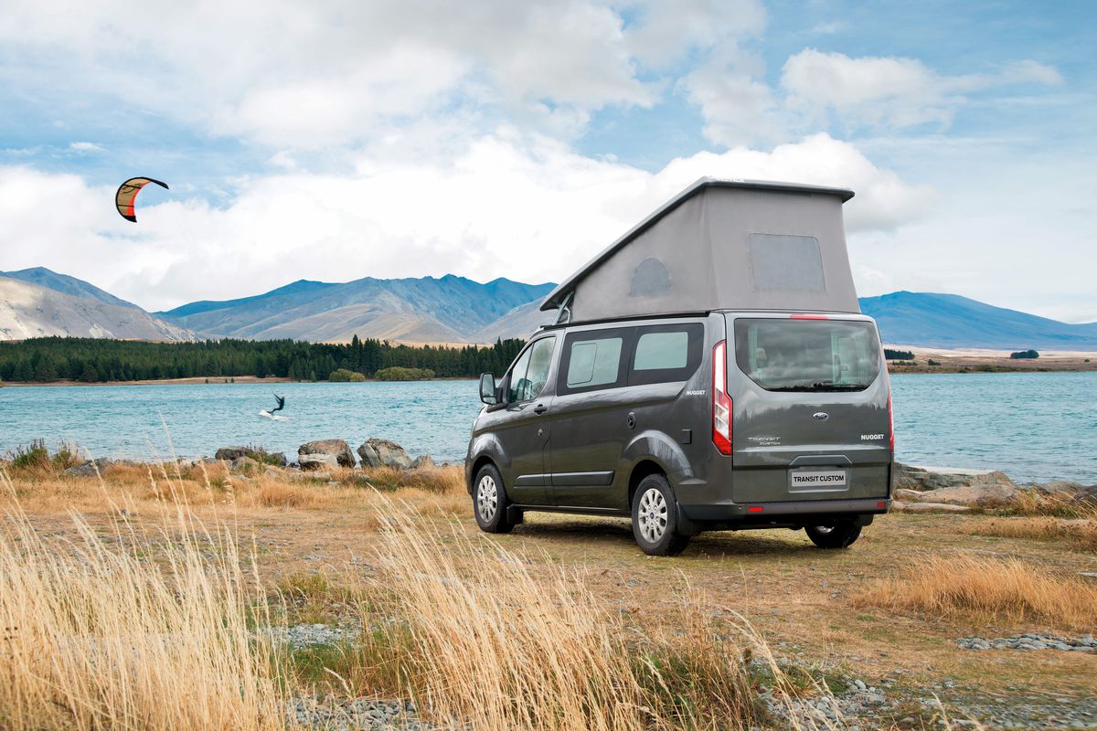A gray pop-top camper van with its roof popped sits in a field next to a mountainous lake. There is a kite boarder on the lake. 