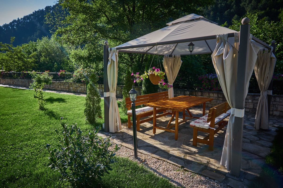 A fabric gazebo stands along in a backyard, equipped with lighting, curtains and a table. 