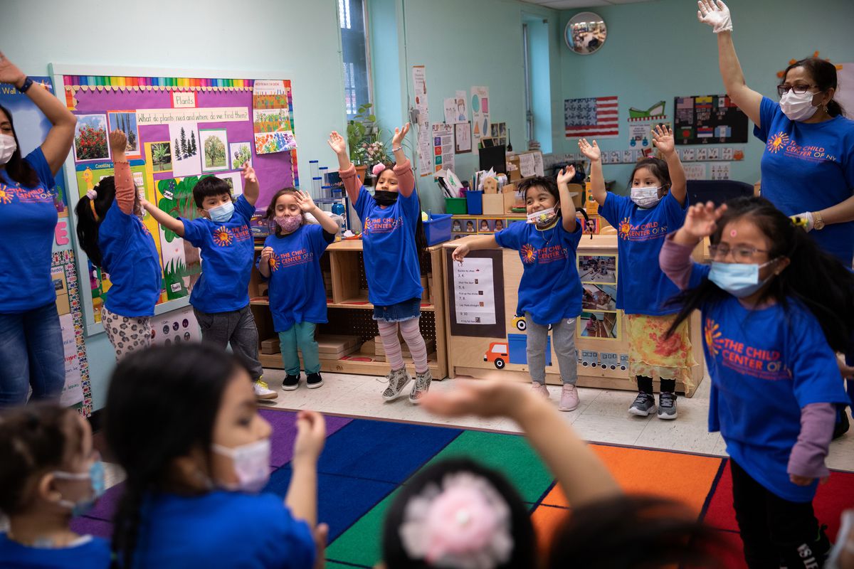 A circle of preschool children in blue T-shirts lift their arms along with their teacher in a group exercise.
