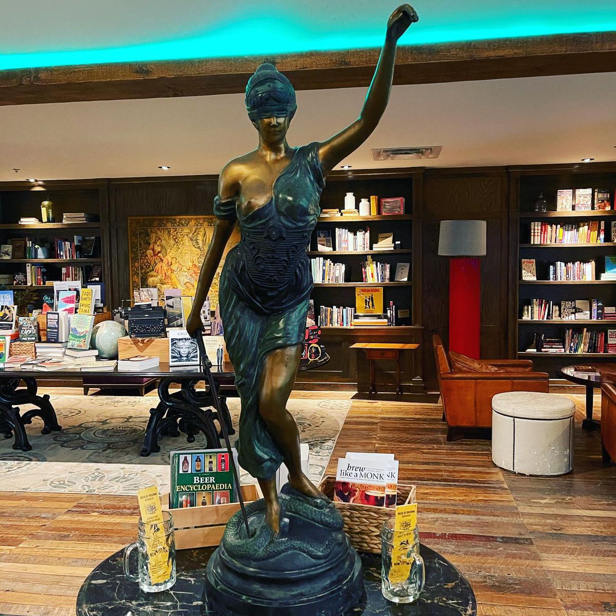 The bookstore on the mezzanine level of Bold Monk Brewing in Underwood Hills, Atlanta, includes titles on beer, cocktail, travel, and cookbook surrounded by dark wooden shelves, leather lounge chairs, plush sofas, and a large bronze statute of Aphrodite as the warrior.