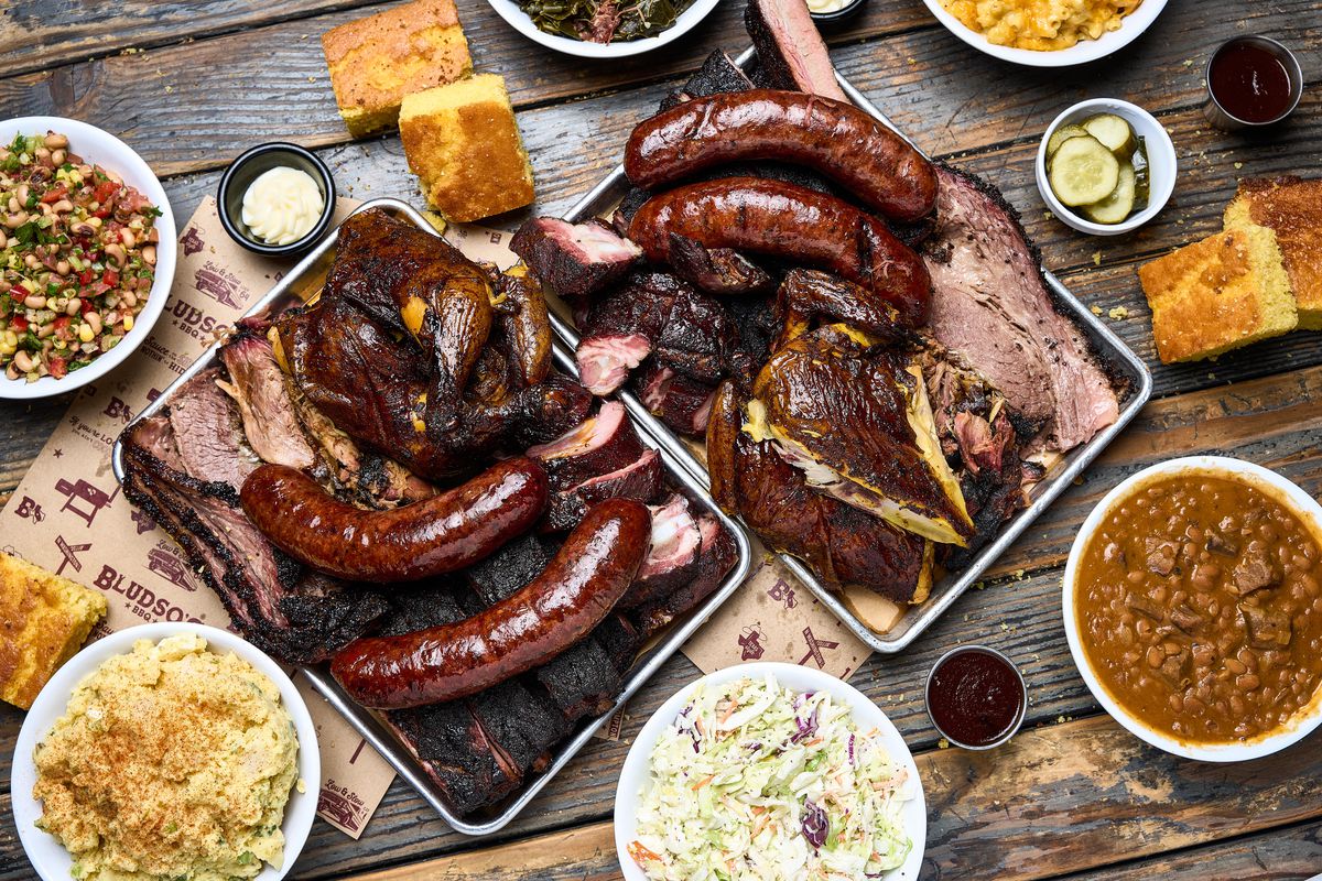 An overhead shot of a wooden table with two metal trays and lots of barbecue, including red hot links, at daytime at a restaurant.