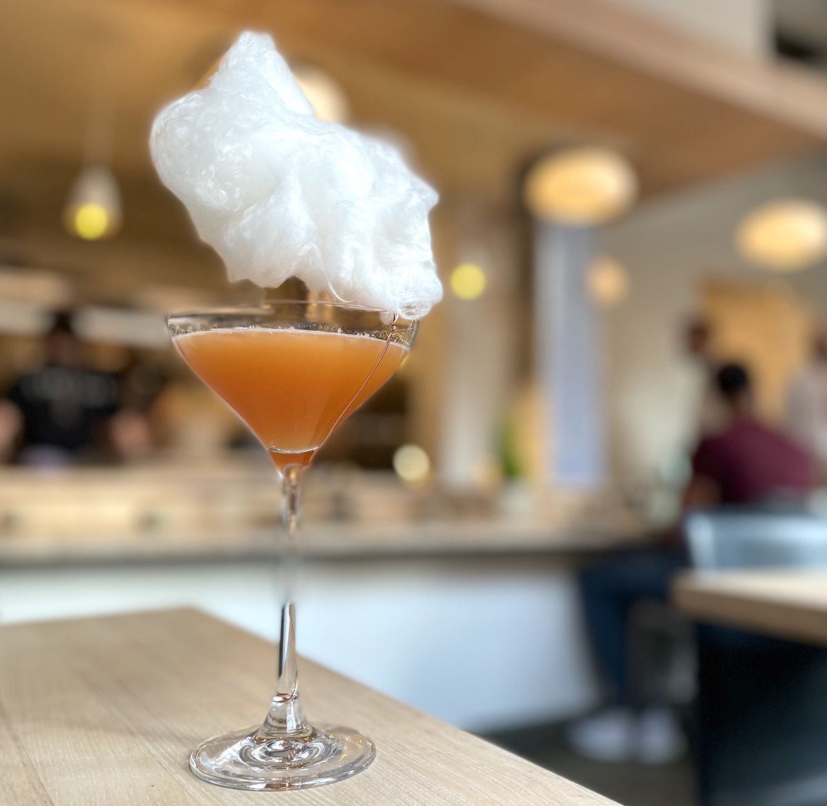 A martini glass is filled with orange liquid on a blonde wood table. It appears that there is a floating cloud above the glass, because a cotton candy puff is placed on it. 