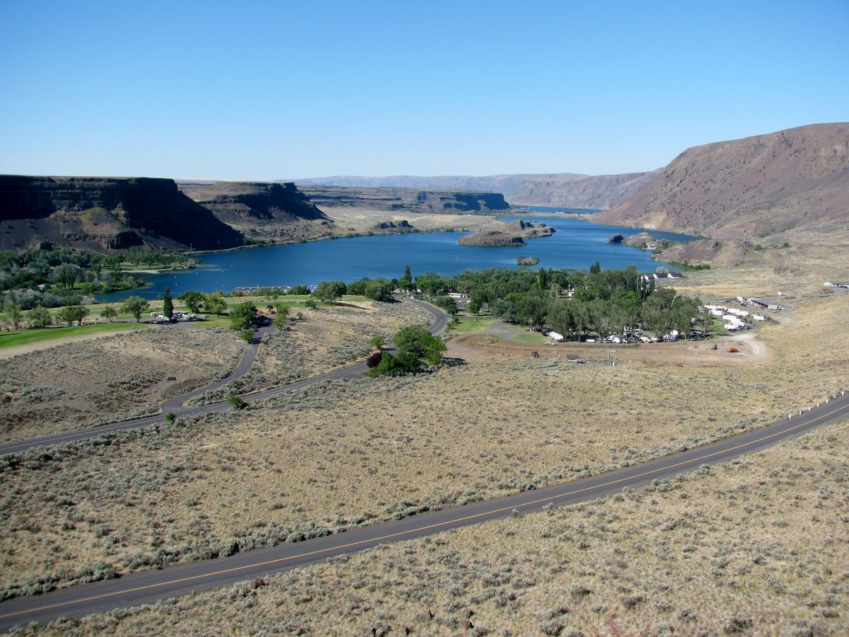 A narrow road runs through a plain of dry glass. A blue lake is surrounded by buttes in the distance, and trees on the side closest to the camera.
