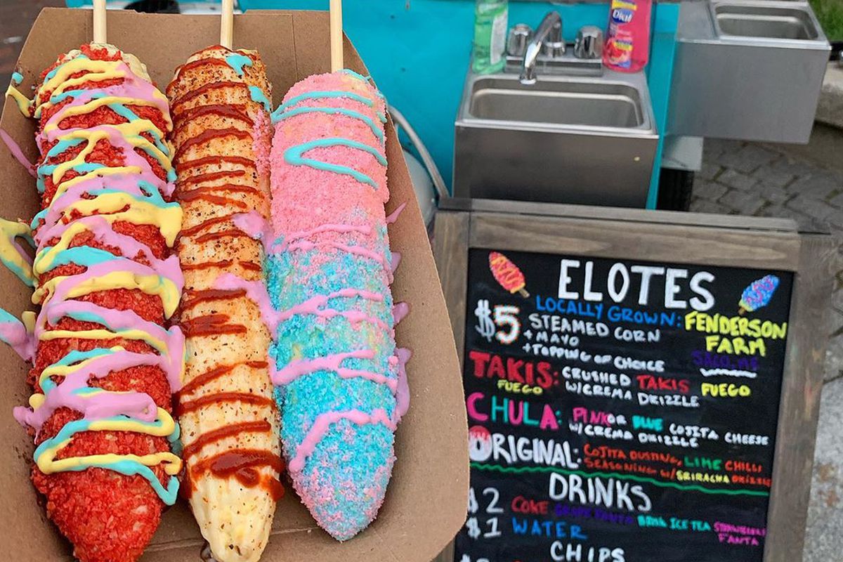 Three ears of corn with bright red, blue, and pink seasoning line a cardboard tray, with a colorful chalkboard sign displaying an elote menu in the rear