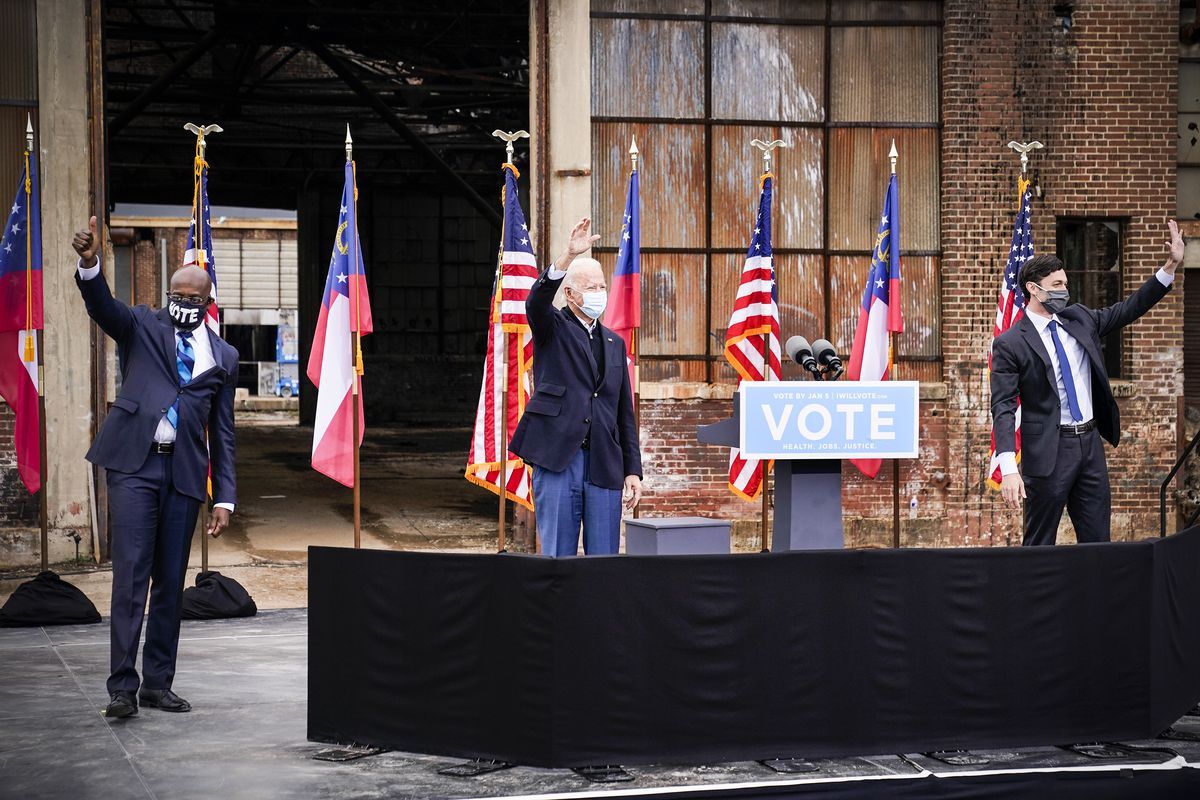 Biden, in a navy sportscoat and blue slacks, waves while standing between — and six feet from — Warnock and Ossoff. All three have on masks; Warnock and Ossoff both have on dark suits with blue ties. The three men are on a stage, outside, set up in front of an industrial building that’s decorated with US and Georgia state flags.