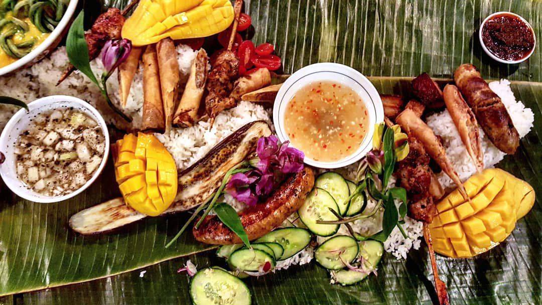 Cut mango, sliced cucumbers, sausages, and other meats form a spread atop banana leaves in a kamayan-style Filipino dinner.