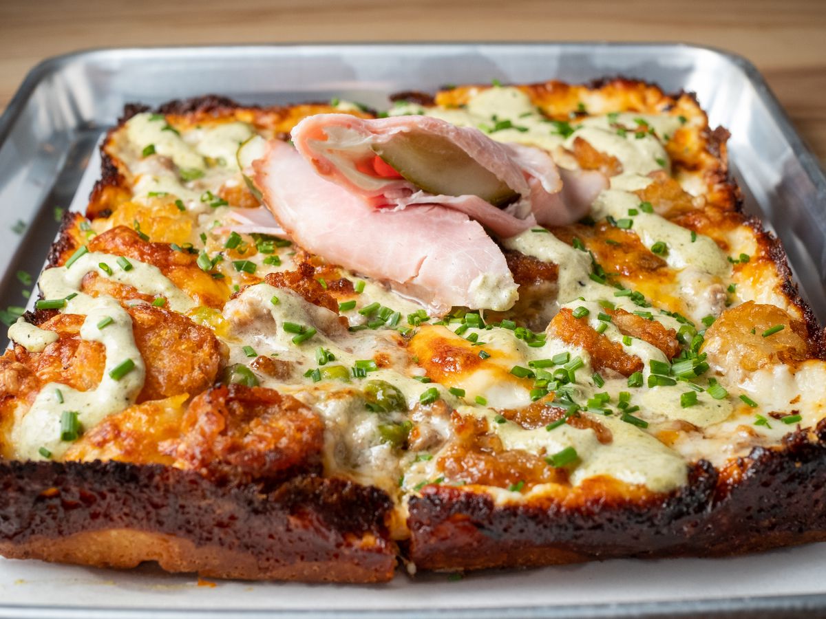 A rectangular pizza topped with tater tots, creamy sauce, an a pickle wrapped in ham garnish