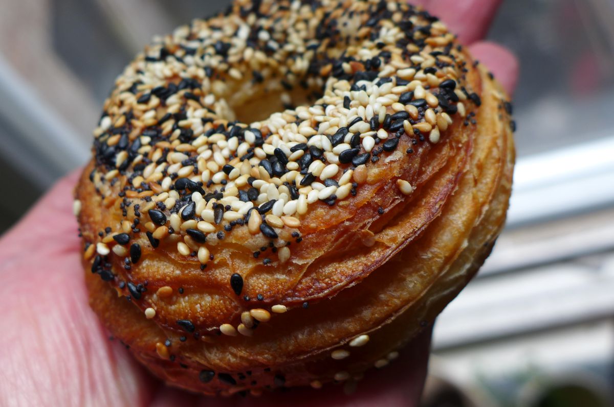 A crusty round pastry held in a hand, with all sorts of seeds and other things scattered on top.