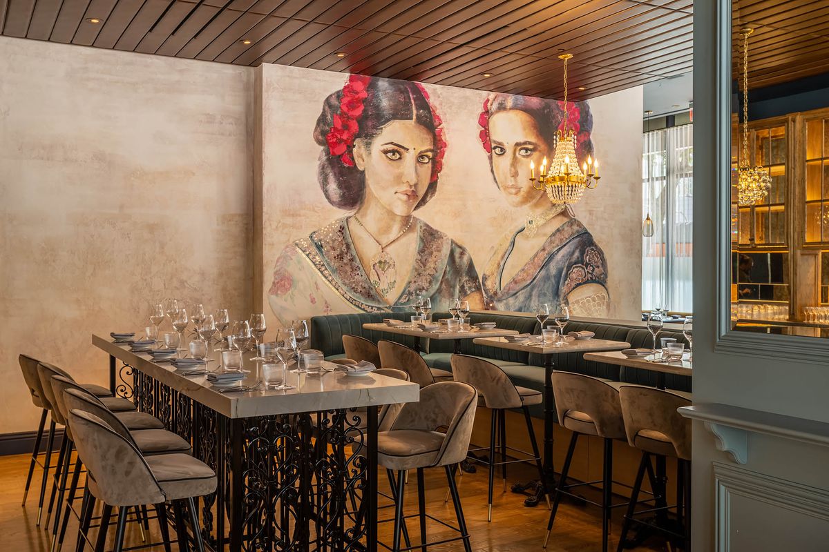 A side look at a mural of two women inside of a tan restaurant at daytime with slate-colored tables and chairs.