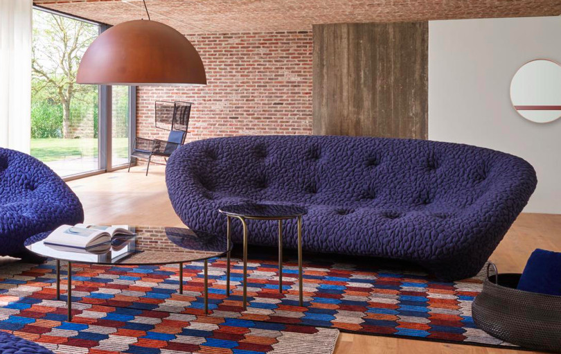 A blue modern couch with a wood lamp over the top of it and a patterned rug beneath. 