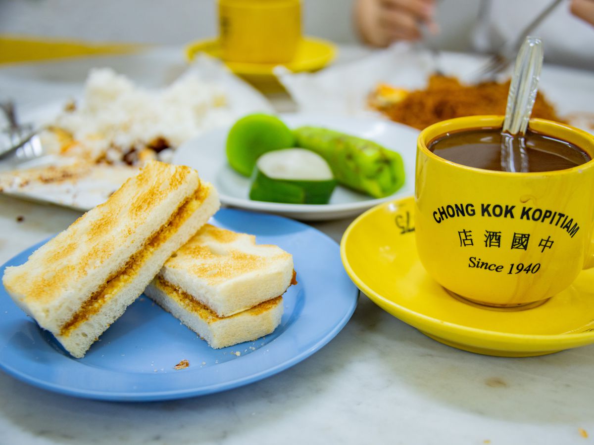 Roti bakar (coconut jam sandwich) sliced in half and stacked on a plate, beside a variety of desserts and a bright mug of coffee bearing the restaurant name