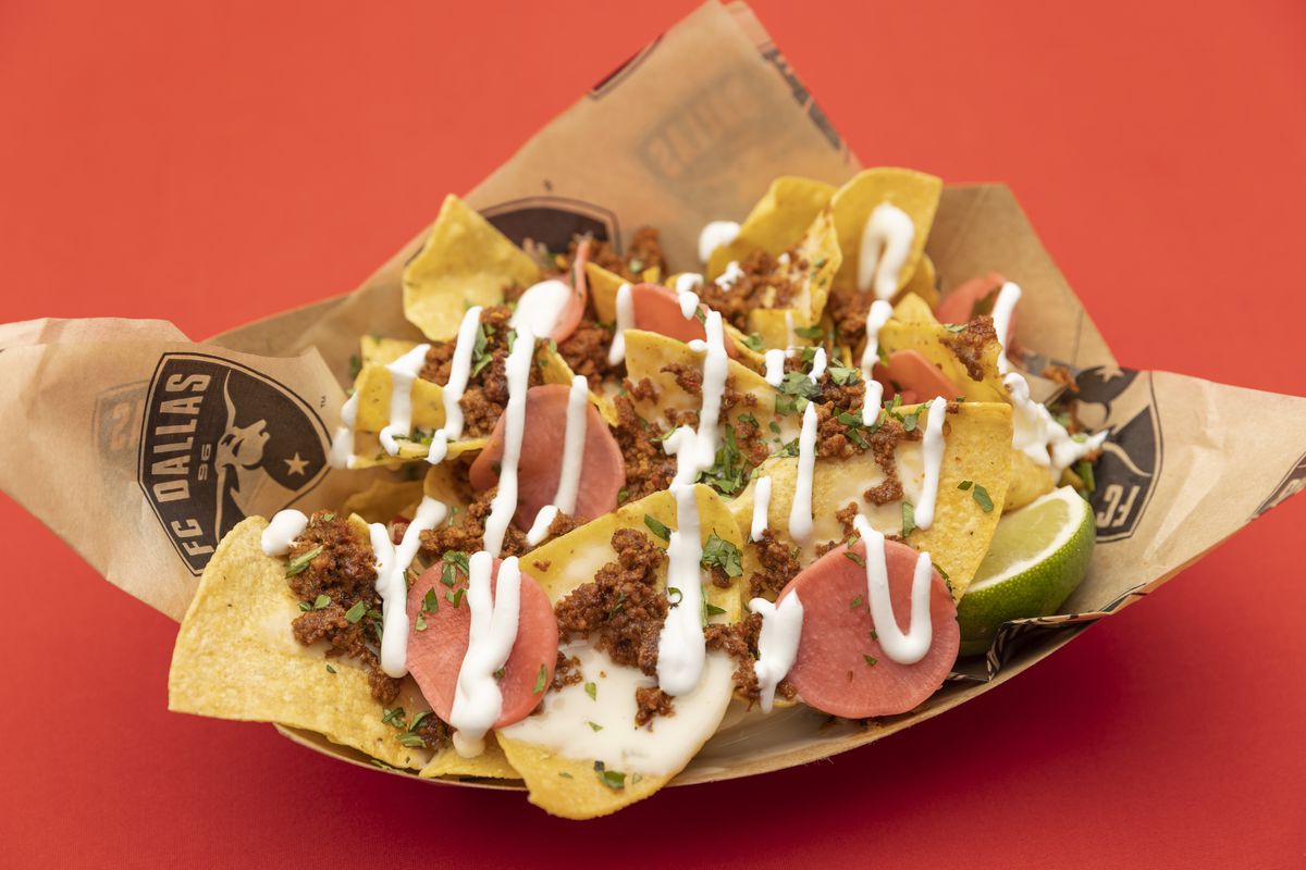 A cardboard container holds chips covered in chorizo, pickled radishes, jalapeno queso, sour cream, fresh cilantro, onions, and tomatoes.