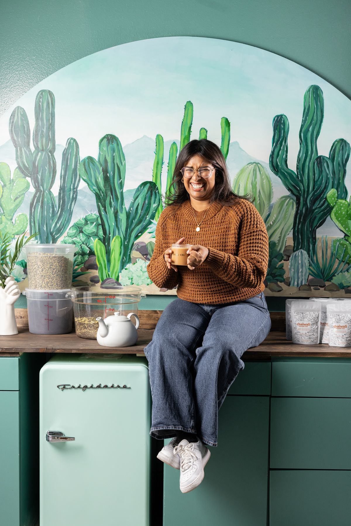 A woman laughs, sitting on a counter top in a kitchen with a cactus mural behind her. In her hands is a mug of chai.