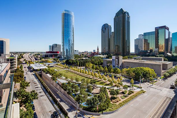 An aerial view of Klyde Warren Park. There is park space in the foreground. In the distance are multiple tall buildings.