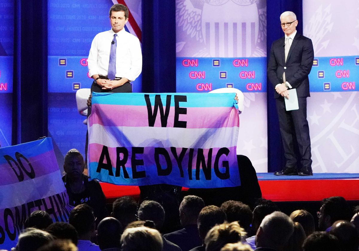 Protester holds up a banner reading “We Are Dying” in front of Pete Buttigieg and Anderson Cooper.