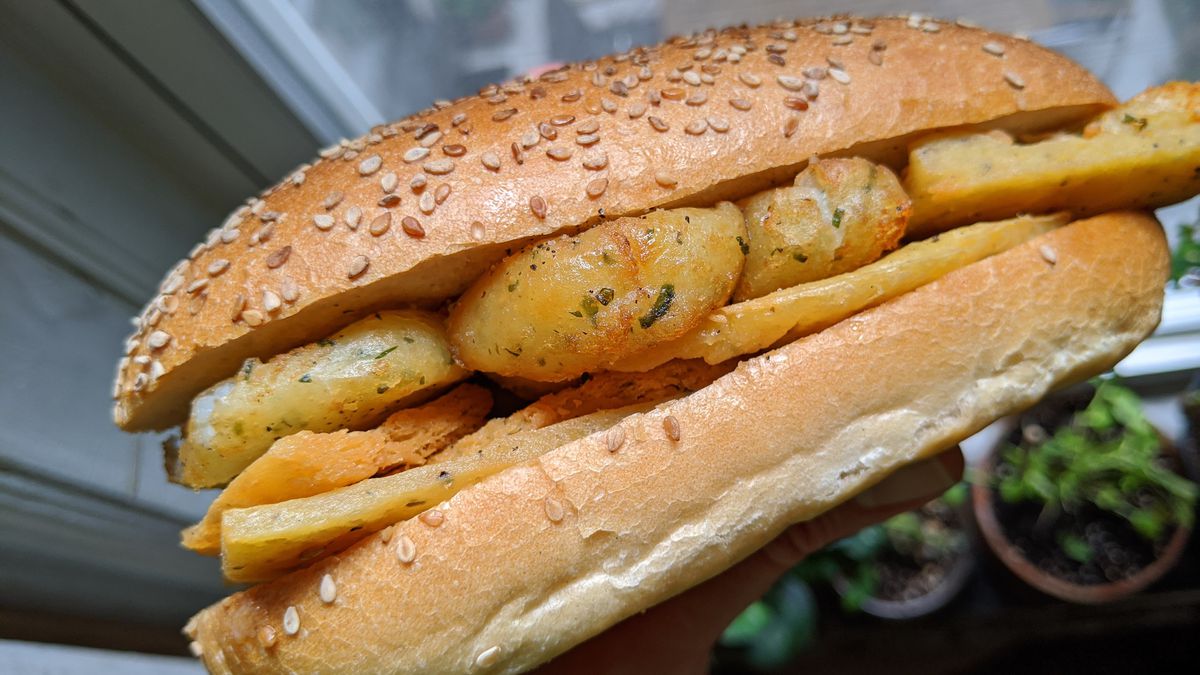 An oblong sandwich with flat browned fritters and round small potatoes.