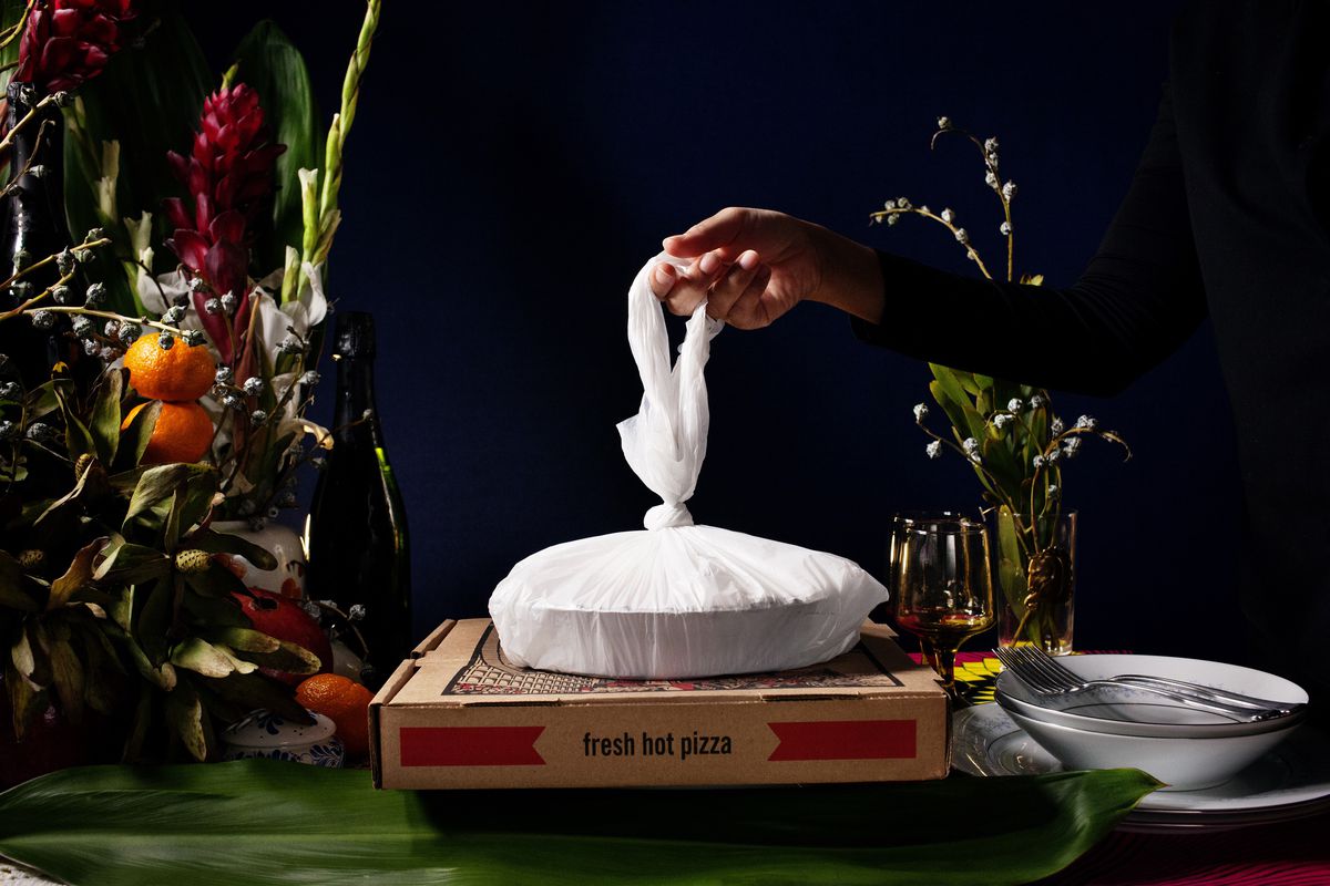 A hand grips a white plastic takeout bag that sits on top of a cardboard pizza to-go box on a table with flowers and greenery in the background