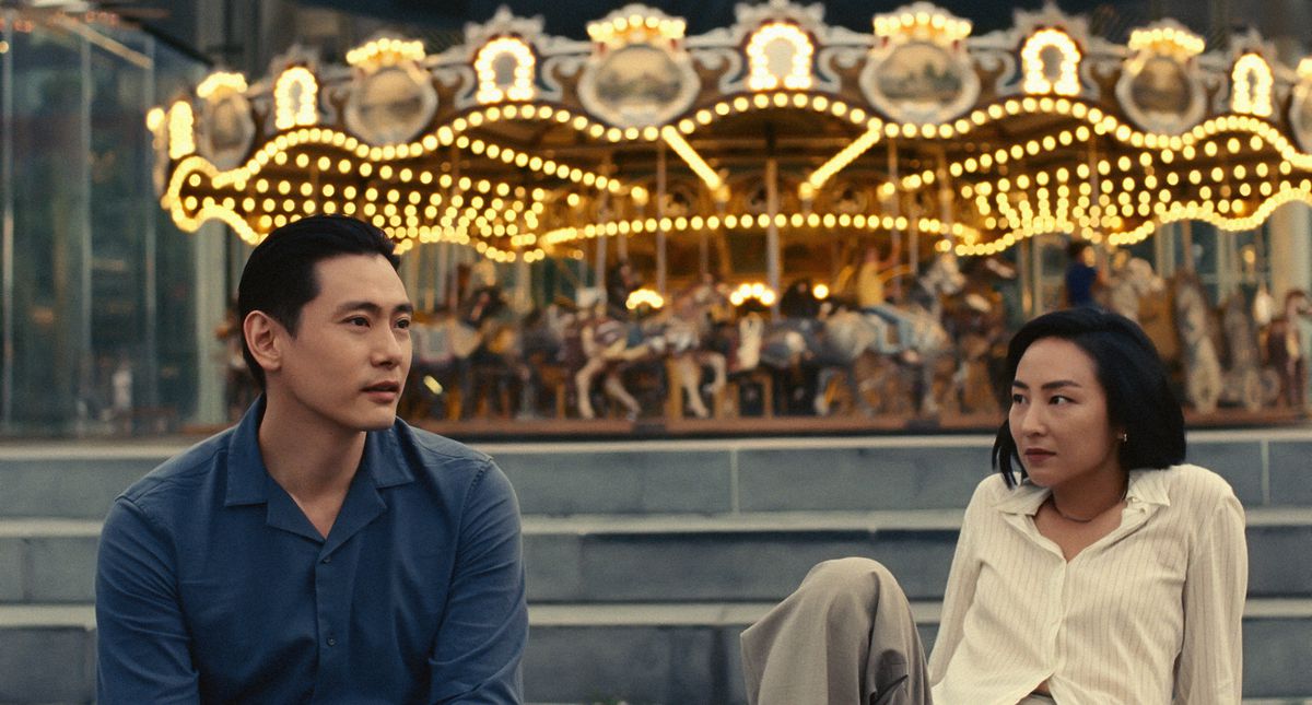 Asian man on the left and Asian women on the right looking at each other, with a carousel in the background.