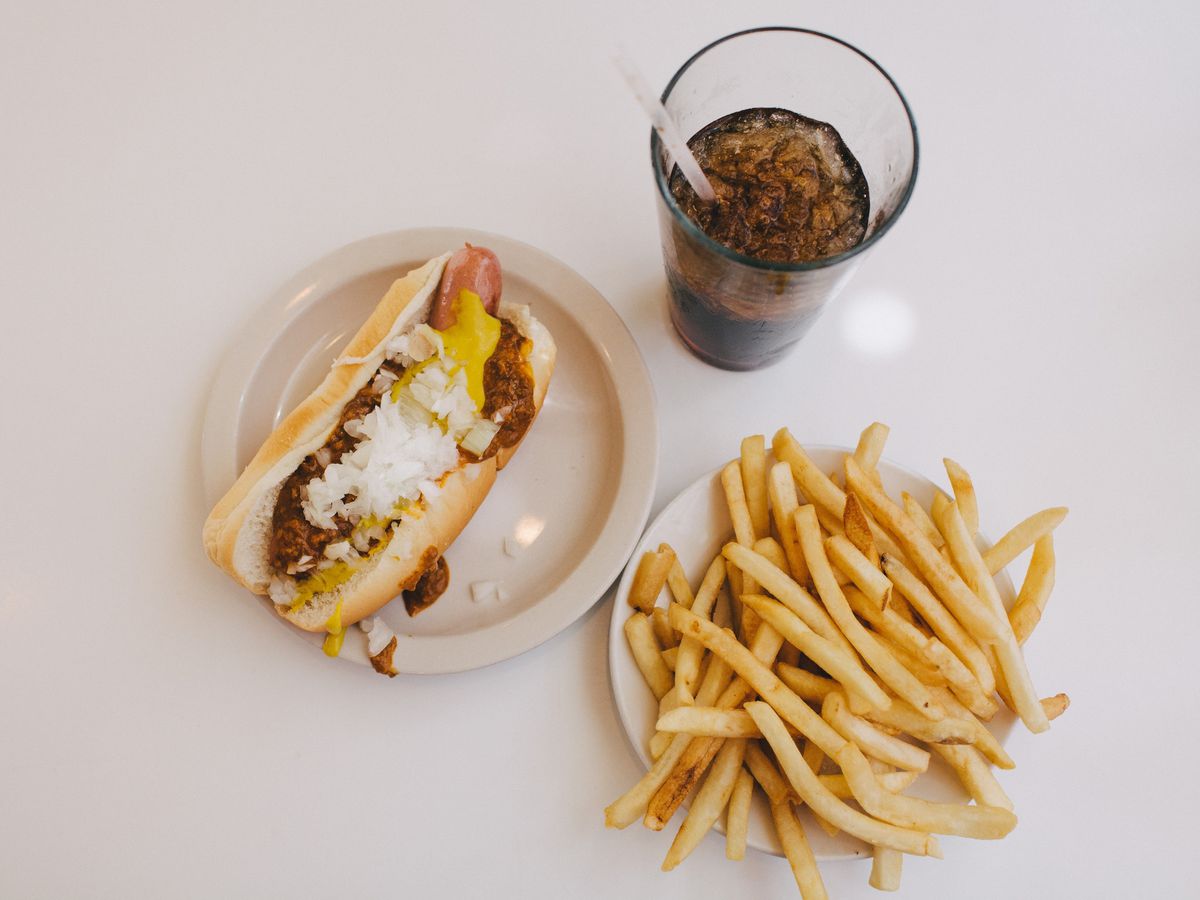A coney dog, a plastic cup of brown cola, and a plate of french fries on a white countertop.