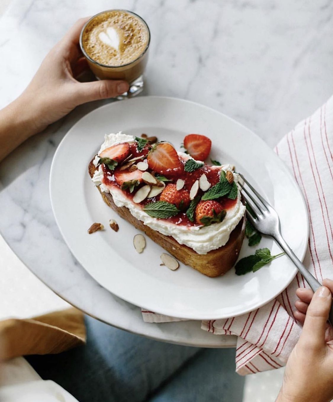 An overhead shop of an open-face tartine with ricotta cheese, strawberries, almonds, and herbs on top. A hand holds an espresso drink with a heart in foam on the left. 