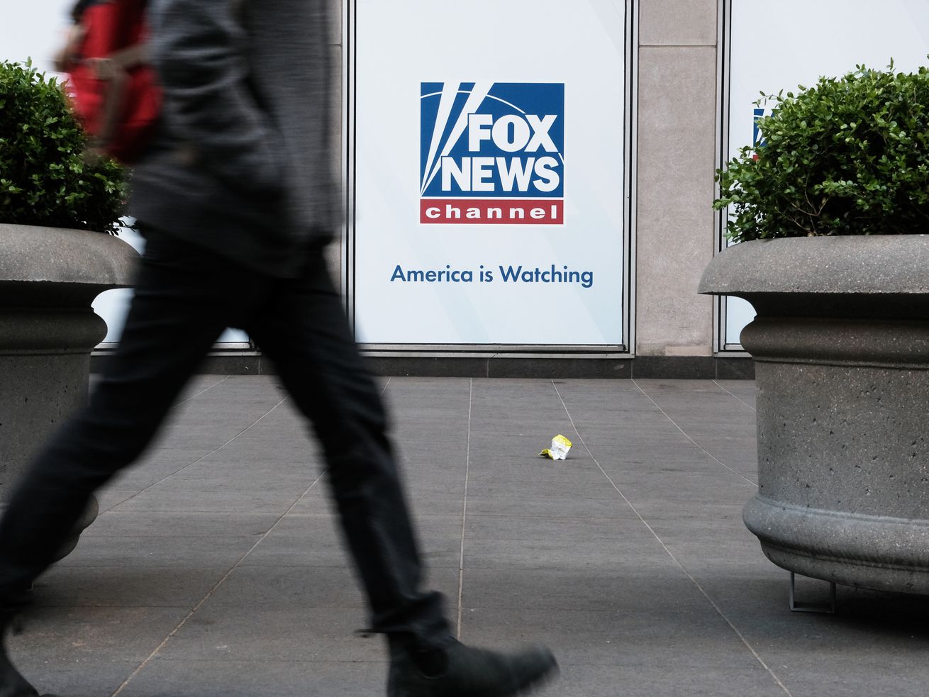 A person walks by the planters outside the News Corp headquarters in New York City, which is displaying a sign that reads “Fox News channel. America is watching.”