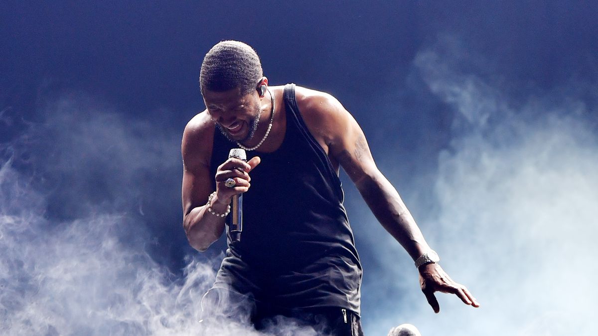 Usher sings into a handheld mic while kneeling onstage, surrounded by smoke.