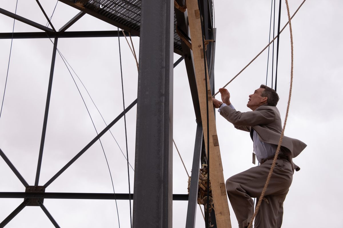 Cillian Murphy as Oppenheimer climbs up the ladder of a tower against a cloudy sky.
