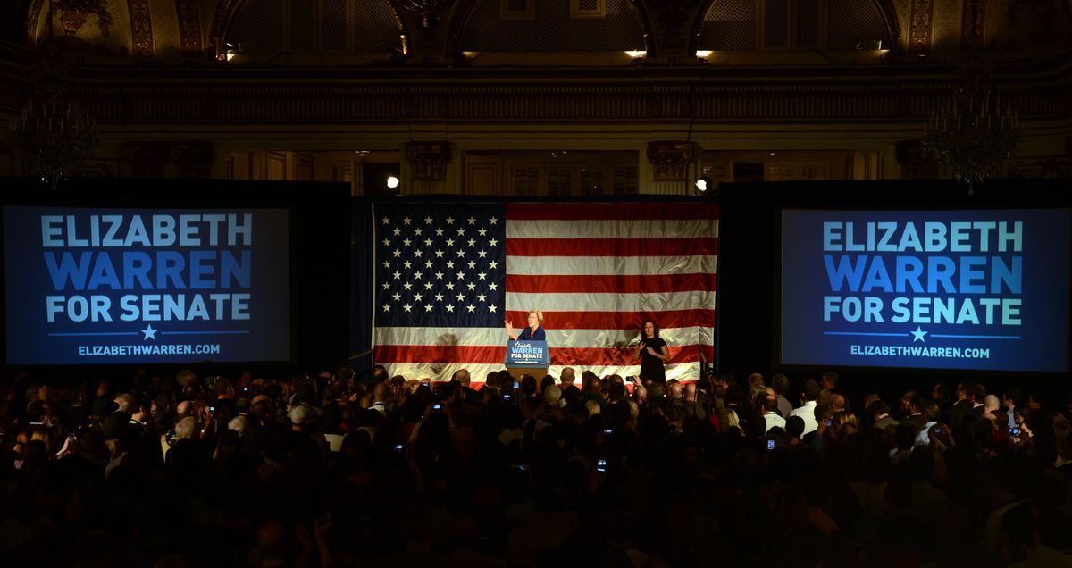 A large crowd at a 2012 campaign event for Elizabeth Warren with her onstage in front of a large American flag and flanked by “Elizabeth Warren for Senate” signs.