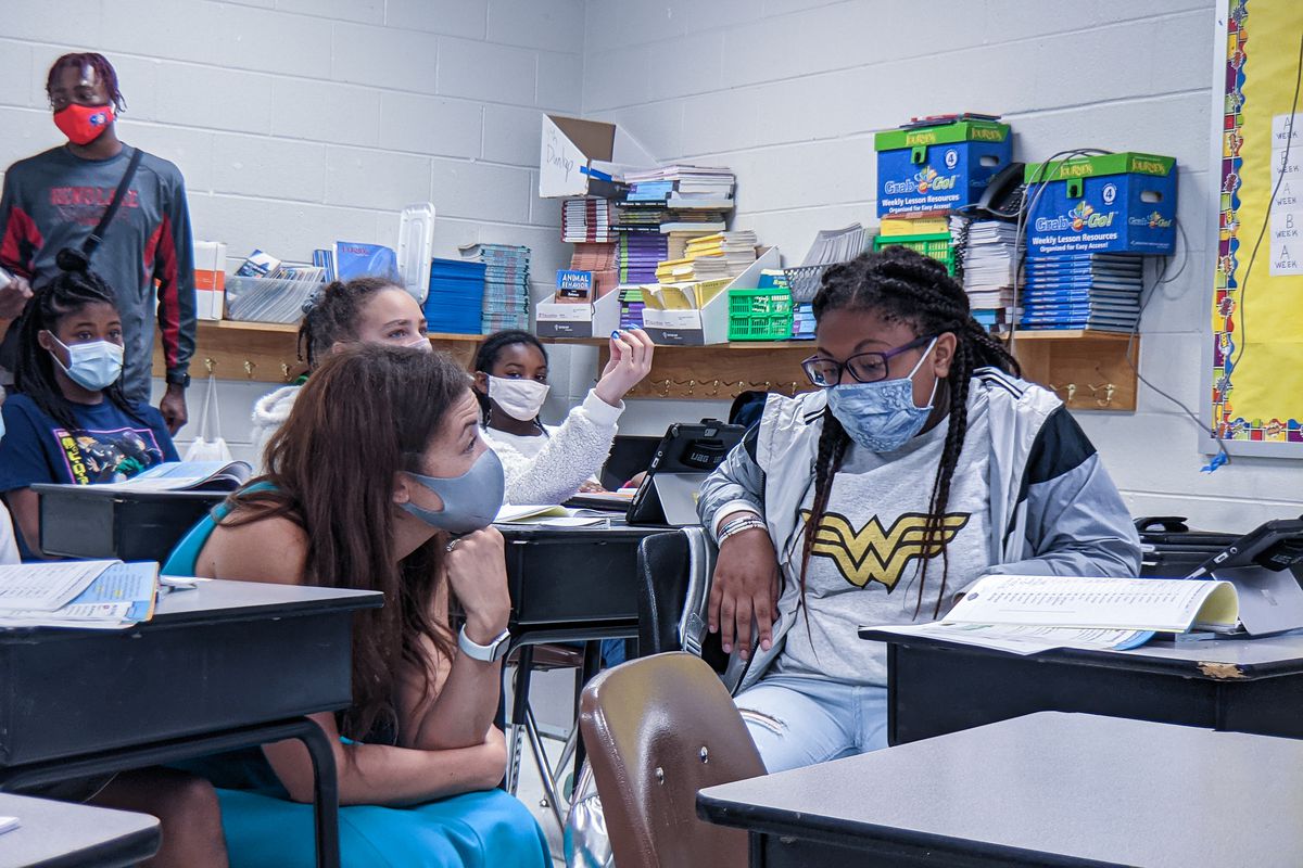 A woman crouches down to speak to a young girl in a classroom. They both are wearing masks.