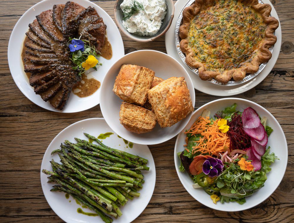 Five round white plates are on a table. Place on them are sliced brisket, grilled asparagus, cheddar biscuits, a colorful salad, and a spinach quiche. 