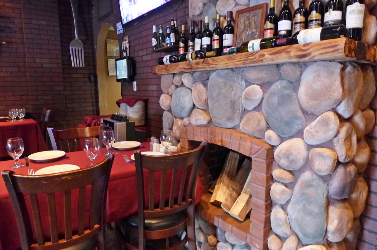 A stone fireplace with a mantle that features bottles of wine lined up, with a table in front with a red tablecloth.