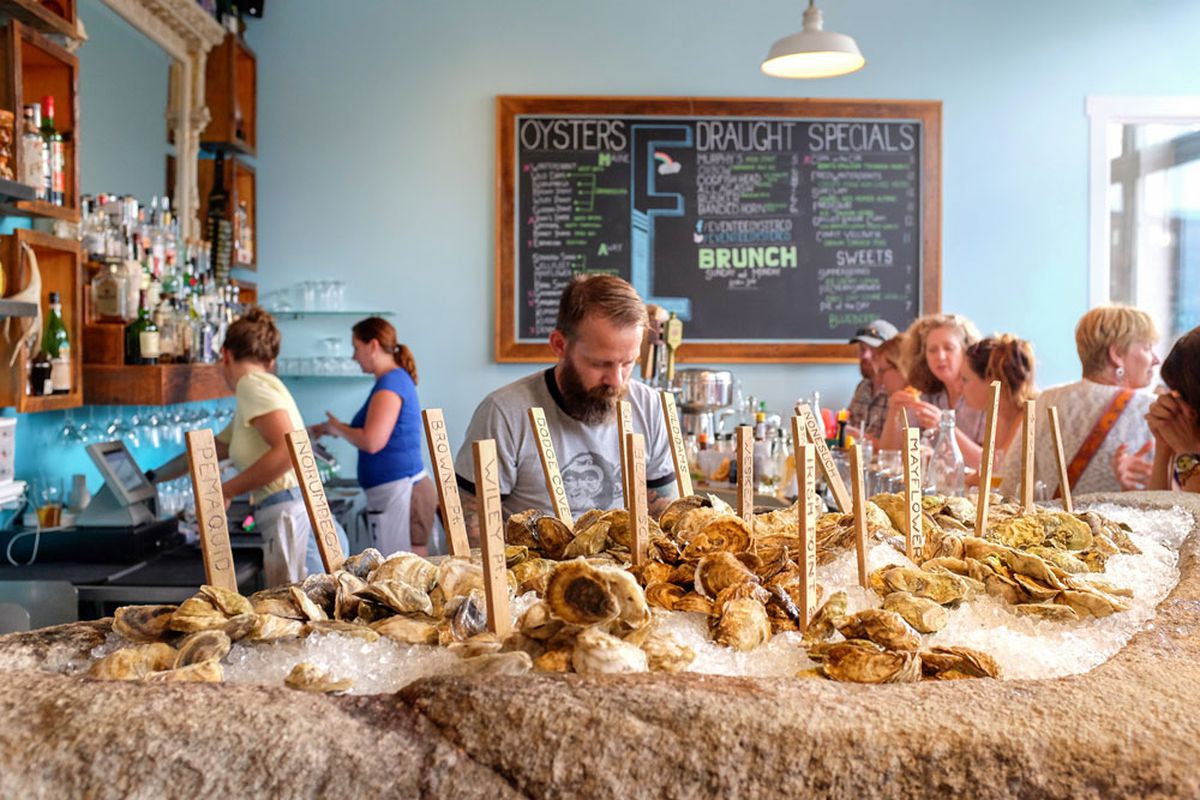 A raw bar full of oysters is in the foreground of the photo, with people working at and dining in a casual restaurant in the background.