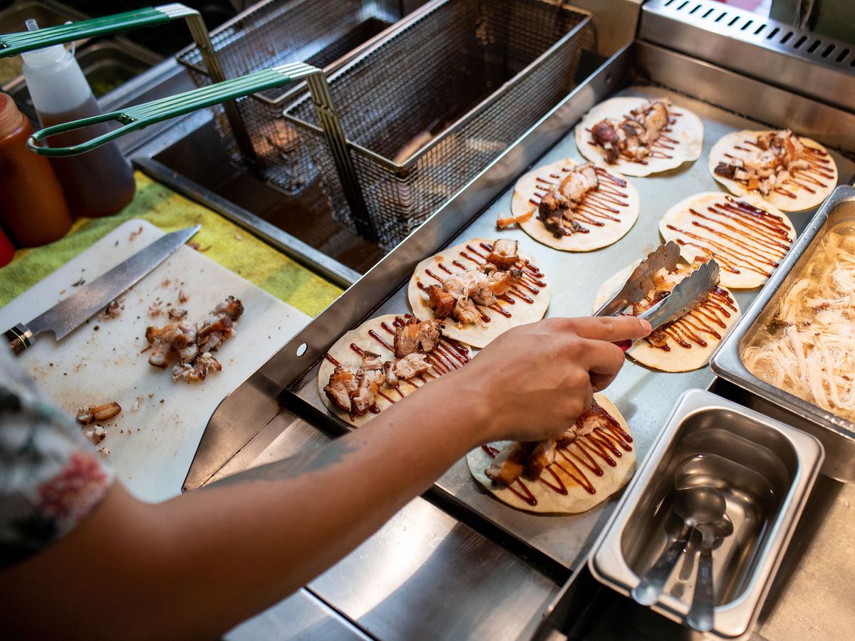 A cook arranges meat with tongs on one of a series of tortillas arranged on a plancha. The tortillas are all already drizzled with sauce. 