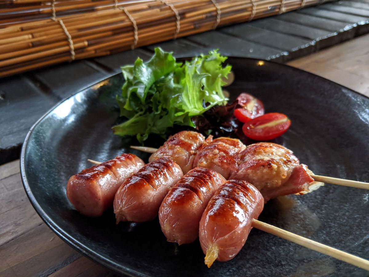 Two wooden skewers of meat on a blue plate —&nbsp;one with pieces of Japanese sausage and one with bacon-wrapped scallops&nbsp;- with a side of greens and sliced cherry tomatoes.