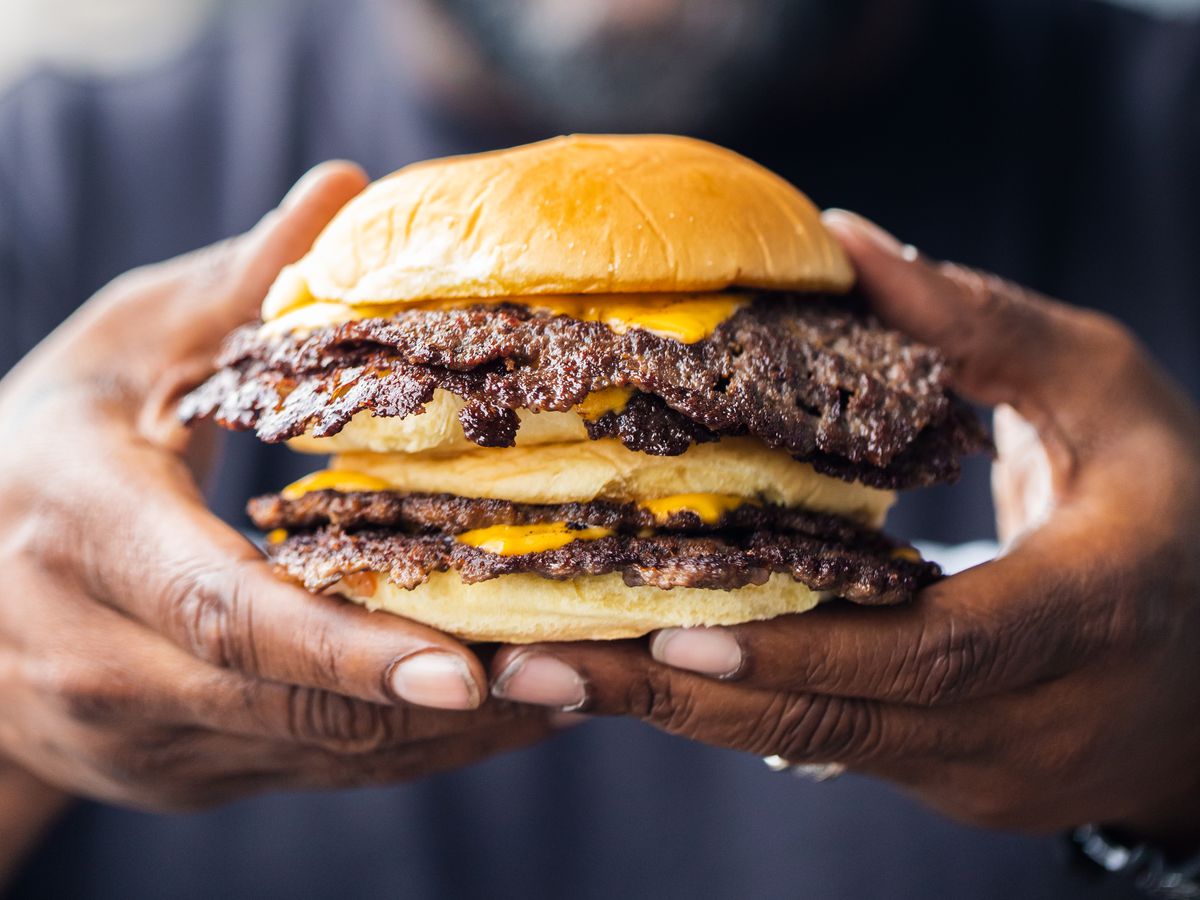 Bun B holding two double-patty Trill Burger smash burgers loaded with cheese.