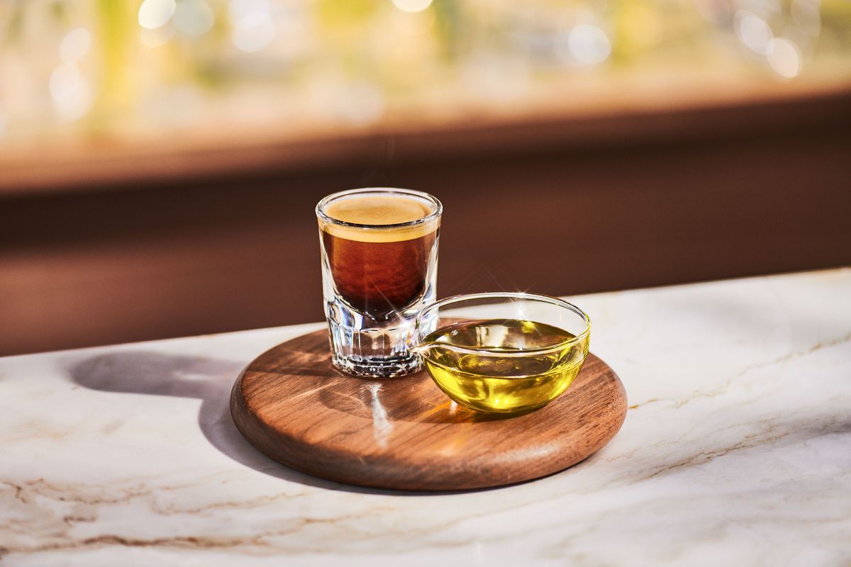 A wooden platter on a marble counter holds a shot of espresso and a bowl of olive oil.