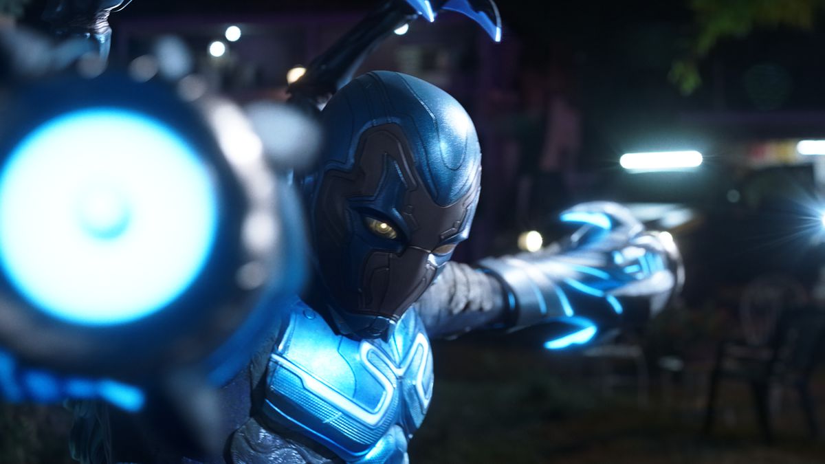 Blue Beetle poses with his arms in the shape of laser canons pointing both directly at and away from the camera in the film Blue Beetle.