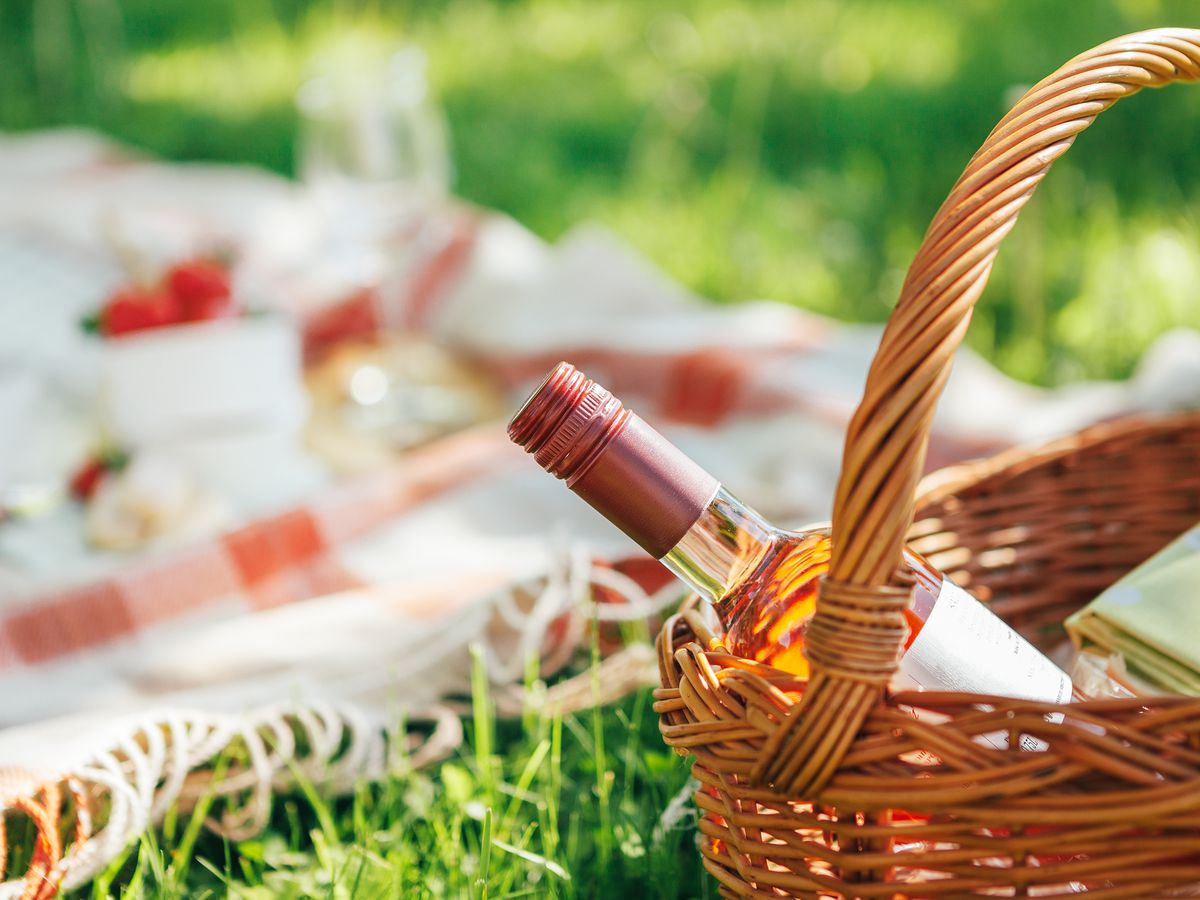 A picnic basket with bottle of rose wine and a checked blanket on the grass with food in the background