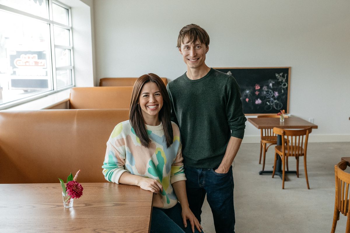 Molly Yeh, wearing a pastel sweater, and Nick Hagen, wearing a grey sweater, stand in Bernie’s restaurant at a brown booth in front of a white wall. 