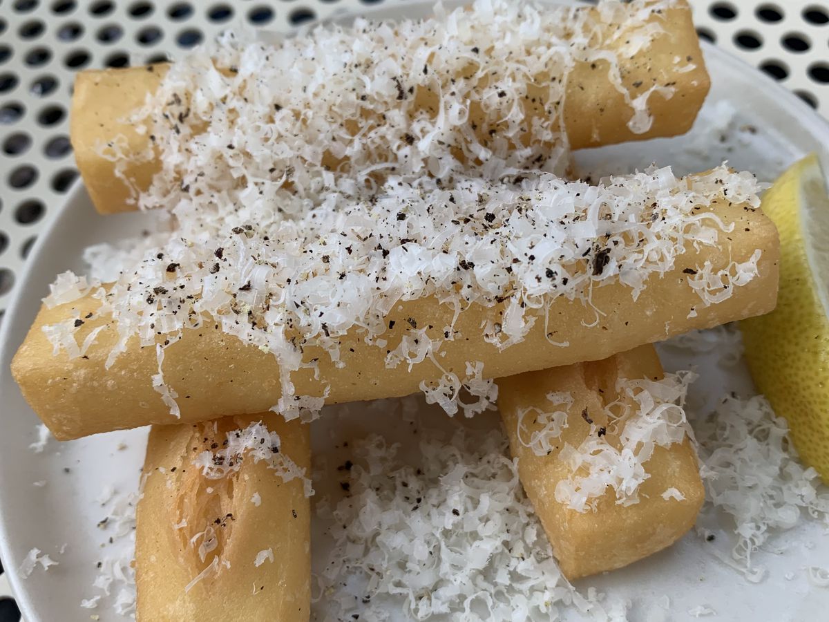 Four lightly tan fried sticks stacked with a pile of fluffy shaved cheese and black pepper on a white plate. 