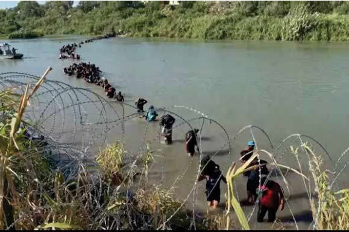 People walking across the river, holding their belongings. Razor wire in the foreground on one side of the river.