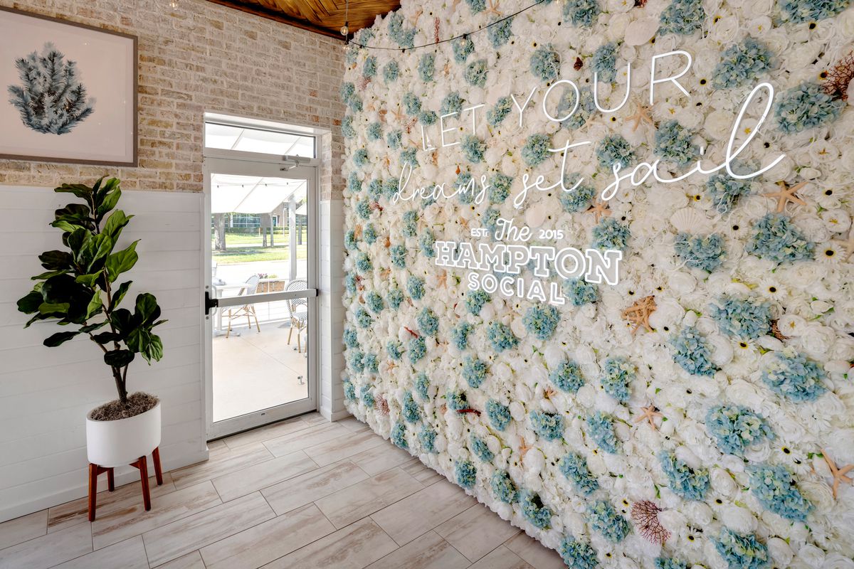 An Instagrammable wall covered in starfish and faux light blue and white flowers includes a sign that reads, “Let your dreams get sail” and the Hampton Social est. 2015. 