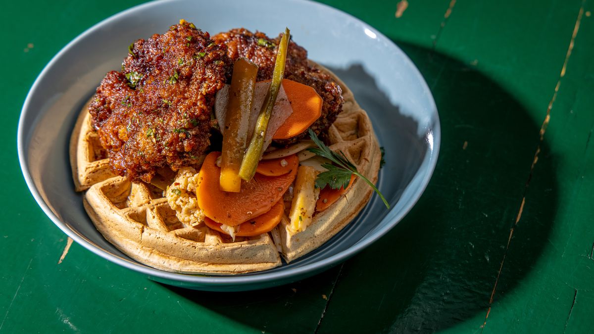 Dark brown sumac fried chicken sits over falafel waffles with pickled carrots; the plate sits above a green table