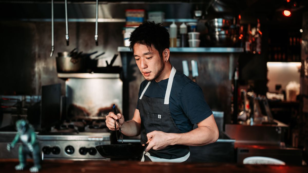 A man with black hair wearing a black t-shirt and an indigo apron holds a wok in one hand and chopsticks in the other. 