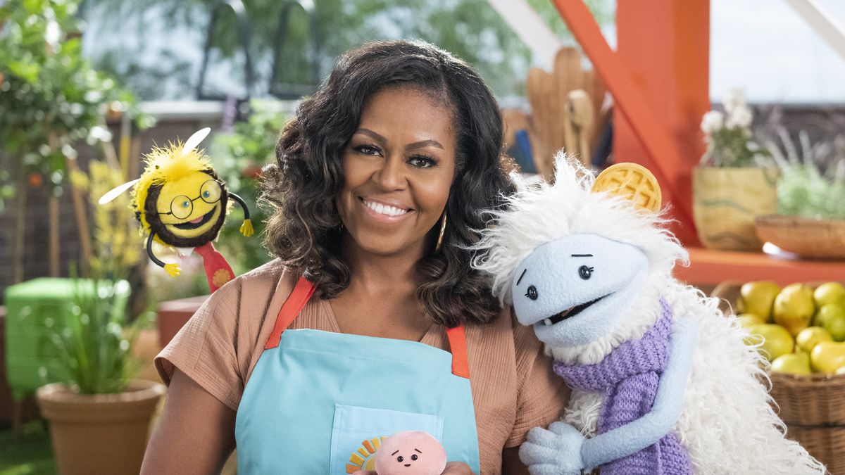 Michelle Obama smiling and surrounded by a puppet bee, a yeti-like puppet, and a little mochi stuffie in her hands.