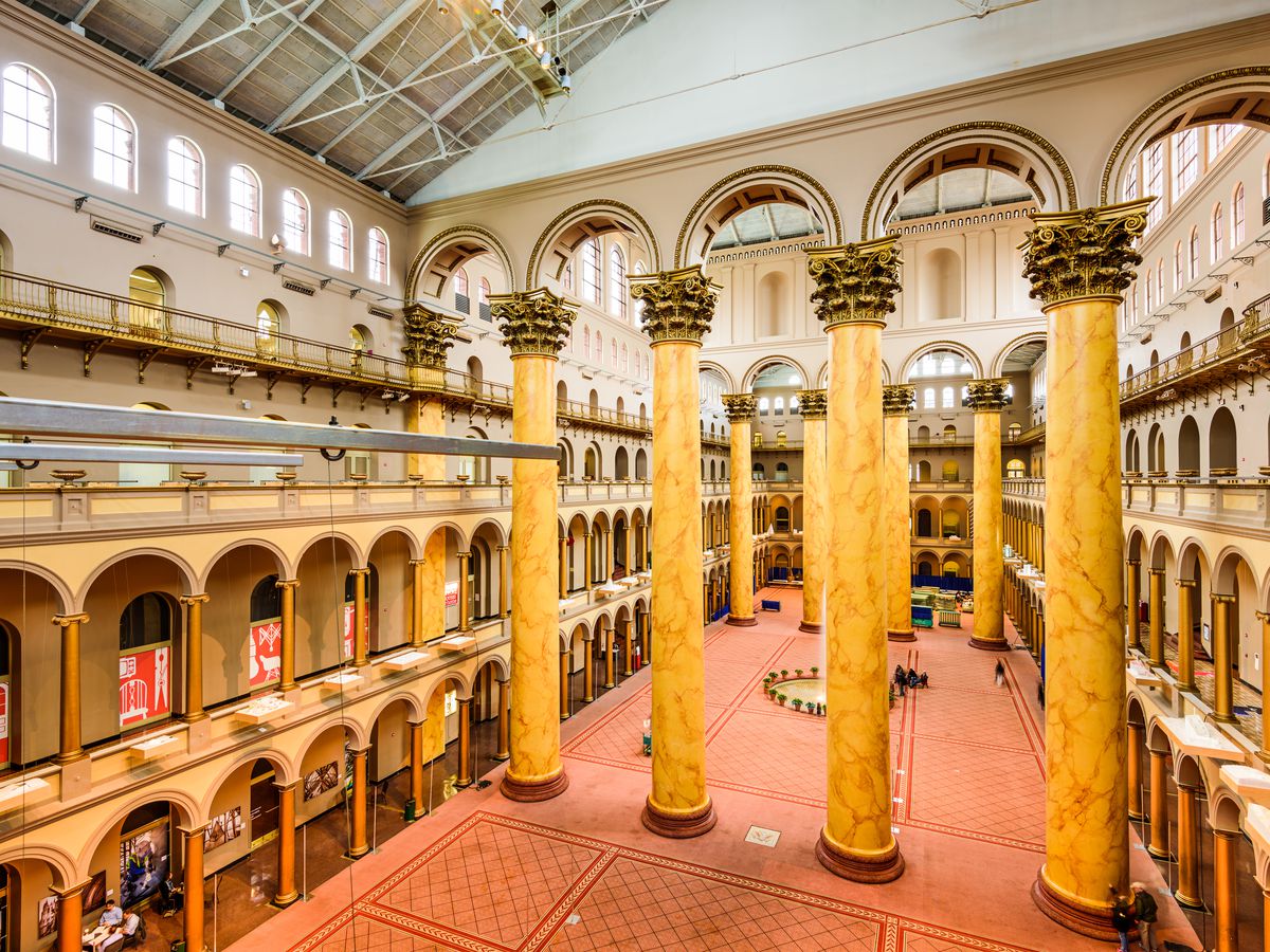 The exterior of the National Building Museum in Washington D.C. The floor is red and there are yellow structural columns. The ceiling is high and arched.