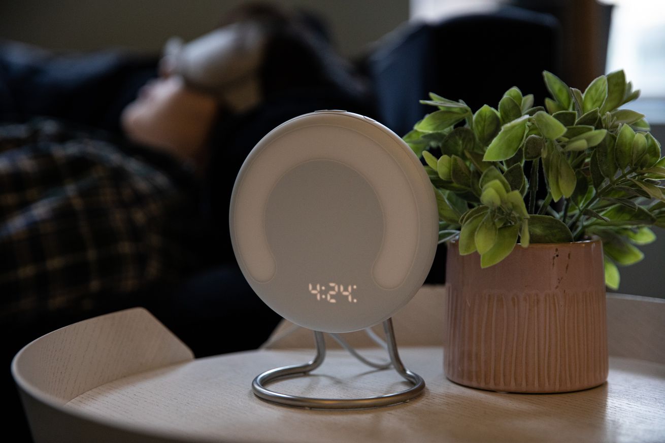 Amazon Halo Rise on a nightstand with a plant, while a person sleeps in the background.