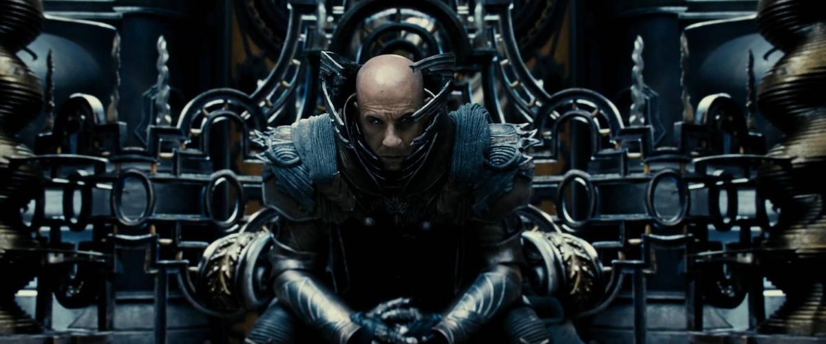 A bald man (Vin Diesel) sits on a throne, dressed in elaborate metallic armor and surrounded by elaborate technology with no clear purpose in The Chronicles of Riddick